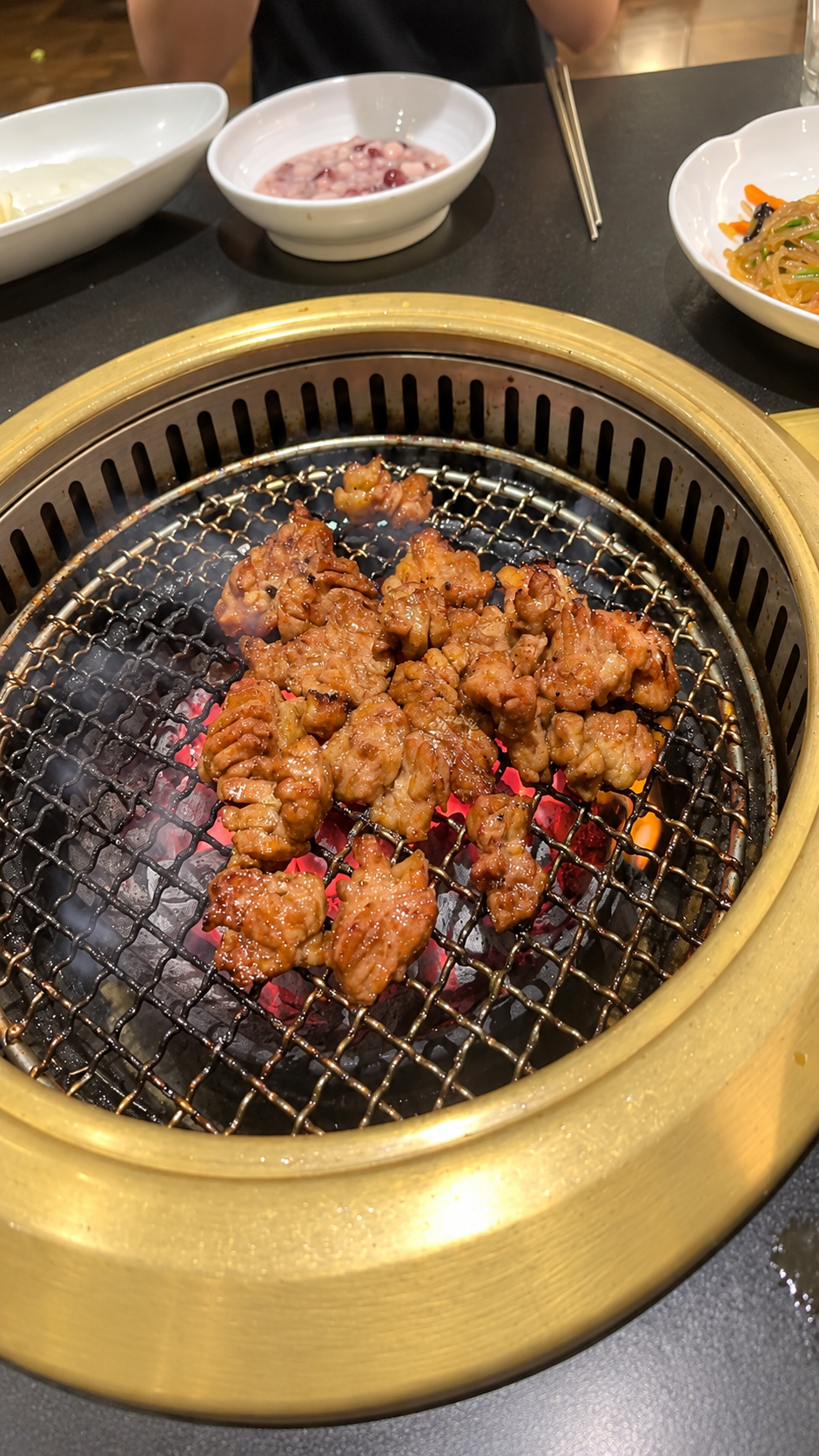 Vertical view of marinated dwaeji-galbi cooking over charcoal with Korean side dishes on the table.